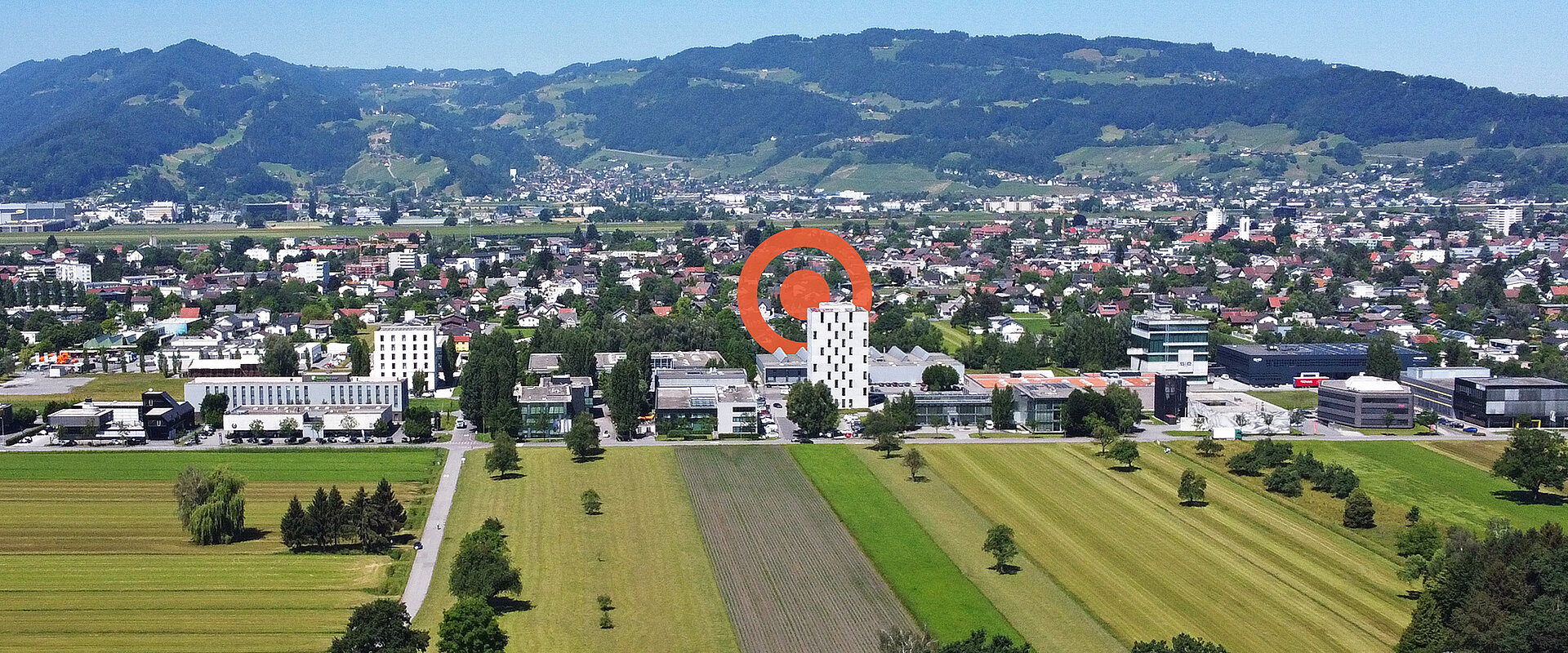 Drohnenaufnahme vom Betriebsgebiet Millennium Park RHEINTAL in Lustenau mit orangem PRISMA Logoelement im Hintergrund.
